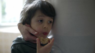 Küçük çocuk annesinin elinden sevgi görürken bir dilim ekmek yiyor. 5 yaşındaki çocuğun, otantik aile sahnesinde abur cubur yerken annesine yaslandığı yakın plan.