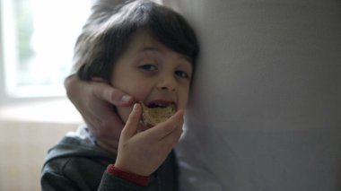 Küçük çocuk annesinin elinden sevgi görürken bir dilim ekmek yiyor. 5 yaşındaki çocuğun, otantik aile sahnesinde abur cubur yerken annesine yaslandığı yakın plan.