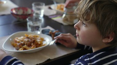 Düşünceli bakışlarla yemek masasında oturan küçük bir çocuğun düşünceli yüzü. Çocuk çatal tutarak uzaklara bakıyor ve yemek yemeyi hayal ediyor.