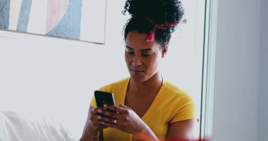 Genç Afrikalı Amerikalı kadın evdeki kanepeye mesaj atıyor. 20 'li yaşlarda Latin kökenli samimi bir kız arkadaşlarıyla internet üzerinden iletişim kurmak için modern teknolojiyi kullanıyor.