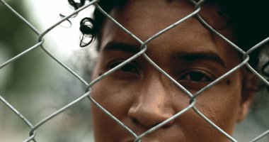 One young black woman trapped behind a fence, close-up hand and face closing eyes in solitude. 20s person struggling with mental illness behind metal barrier depicting depression clipart