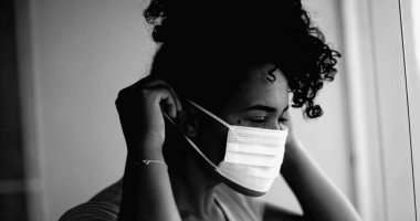 Genç siyahi bir kadın, yoğun dramatik monokromatik portresiyle evde takılıp kalan siyah maskesini takıyor. Daireden pencereye bakıyor.