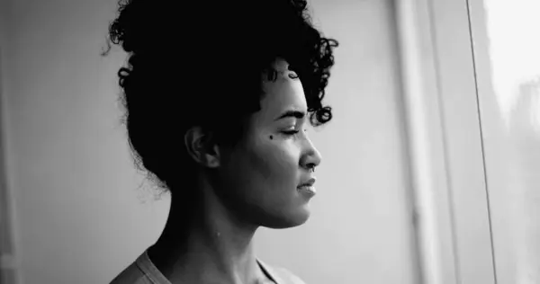 One contemplative young black woman standing by window gazing in the distance with meditative emotion in dramatic monochromatic, black and white. Profile close-up face