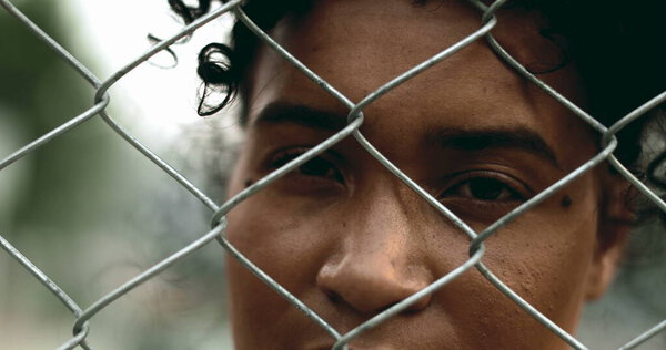 One young black woman trapped behind a fence, close-up hand and face closing eyes in solitude. 20s person struggling with mental illness behind metal barrier depicting depression