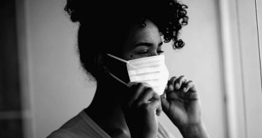 Genç siyahi bir kadın, yoğun dramatik monokromatik portresiyle evde takılıp kalan siyah maskesini takıyor. Daireden pencereye bakıyor.