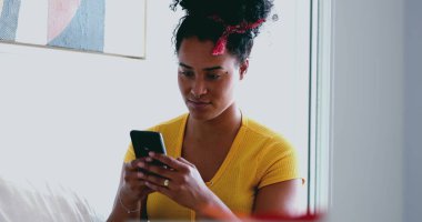 Genç Afrikalı Amerikalı kadın evdeki kanepeye mesaj atıyor. 20 'li yaşlarda Latin kökenli samimi bir kız arkadaşlarıyla internet üzerinden iletişim kurmak için modern teknolojiyi kullanıyor.