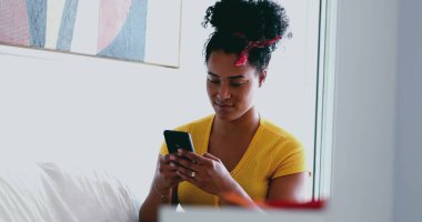 Genç Afrikalı Amerikalı kadın evdeki kanepeye mesaj atıyor. 20 'li yaşlarda Latin kökenli samimi bir kız arkadaşlarıyla internet üzerinden iletişim kurmak için modern teknolojiyi kullanıyor.