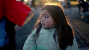 İçsel olarak yalnız 8 yaşında bir kız çocuğu, gün batımında arka planda, parktaki bankta oturmuş, etrafındaki çocukları seyrediyor.