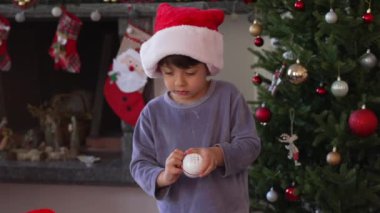 Noel sabahı aile fertleriyle çevrili Noel şapkası takan genç bir çocuk aile hediyelerini açıyor. Paketleri açıyor.