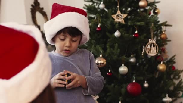 赤いクリスマスの帽子とパジャマを身に着けているかわいい少年は 家族に囲まれた朝にプレゼントを開きます — ストック動画