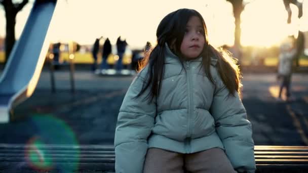 平和な8歳の少女が冬季の日没時に周囲を観察する公園のベンチに座った — ストック動画