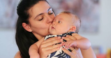 Candid young mother holding month old baby kissing and listening to conversation