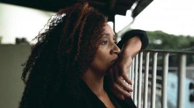 Üzgün, orta yaşlı, düşünceli bir kadın ev balkonundan şehir manzarasına bakarken derin bir üzüntü ve yalnızlık hisseden melankolik bir ifadeyle 50 'lerin Afrika kökenli