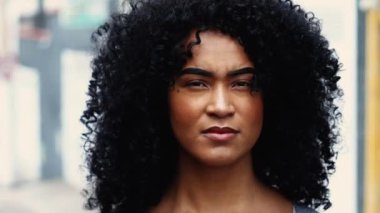 Kendine güvenen, ciddi, kıvırcık saçlı, şehir ortamında duran siyah bir kadının portresi. Güney Amerika Latin Kişi sokağın ortasında sert bir ifadeyle kameraya bakıyor.