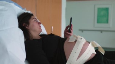 Hamile kadın cep telefonu cihazına bakıyor. Doğum öncesi kasılmalar sırasında karnında ultrason kayışlarıyla hastanede bekliyor.