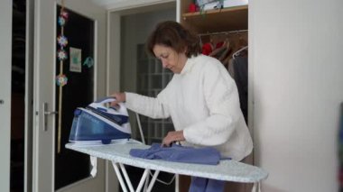 Kıdemli kadın, günlük rutin yaşam tarzıyla kıyafetleri ütülüyor. Evde ev işleriyle ilgilenen yaşlı bir kadın.