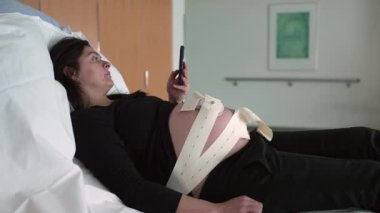 Hamile kadın erken doğum sırasında fetüsün kalp atışlarını ölçmek için ultrason kayışlarıyla hastanede yatarken telefonu kontrol ediyor.