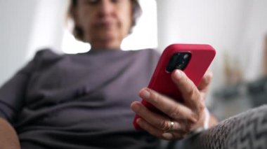 Ev koltuğunda oturan üst düzey bir kadının cep telefonu cihazının yakın çekimi, içeriği internetten okuyan kişi, teknoloji telefonu.