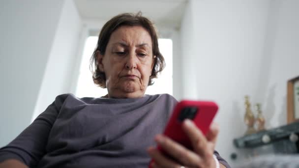リビングルームでソファーに座っている間 オンラインでコンテンツを読む携帯電話デバイスを保持するシニア女性 画面を見ている70代の深刻な表現 — ストック動画