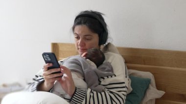 Anne yeni doğan bebek göğsüne yaslanırken cep telefonu kullanıyor, kadın kulaklık takarak yatağa uzanıyor, bebek bebekle birden fazla iş yaparken telefon ekranına bakarak mesaj yazıyor.