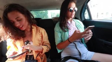 İki genç kadın arabanın arkasında akıllı telefon kullanıyor.