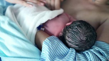Yenidoğan 'ın İlk Beslenmesi- Hemşire bebeğini battaniyeyle örterken, anne de hastane kliniğinde cilt cilde teması vurguluyor.