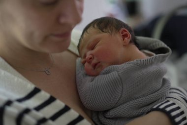 Yeni doğmuş bebeğini sıcacık gri bir kazağa sarılmış bir annenin yakın, samimi bir fotoğrafı. Bebek mışıl mışıl uyuyor, derin bir anne sevgisi ve bağlantı anı yakalıyor, sıcaklık ve şefkat yayıyor..