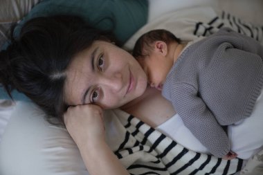 Bir anne ve yeni doğmuş bebeğinin birlikte yattığı ve bebeğin annesinin göğsünde dinlendiği hassas bir sahne. İkisi de rahat ve güçlü bir bağ sergiliyorlar, bebek rahat gri bir kazak içinde, sıcaklık ve sevgiyi yansıtıyor..