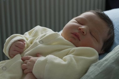 Yeni doğmuş bir bebeğin, yumuşak sarı bir tulumun içinde huzurlu bir şekilde uyuduğu, küçük ellerinin nazikçe dinlendiği yakın plan bir fotoğraf. Sahne, bebeğin narin ve huzurlu doğasını vurgular..