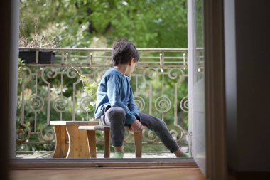 Genç bir çocuk, mavi uzun kollu gömlek ve gri pantolon giymiş bir balkondaki küçük ahşap bir bankta oturuyor. Derin düşüncelere dalmış gibi görünüyor, yere bakıyor, sakin bir yeşillik zeminiyle..