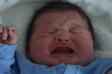 Yeni doğmuş bir bebeğin ağlayan yüzünün yakın çekimi, saf duygularını ve mavi tulum giymiş minik ellerini gösteriyor, yeni hayatın hassasiyetini vurguluyor.. 