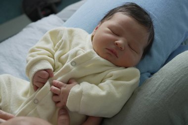 Küçük bir el göğsünde dinlenirken, sarı tulumun içinde huzur içinde uyuyan yeni doğmuş bir bebeğin yakın çekimi. Sahne sakin ve narin..