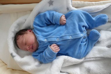 Mavi tulumlu yeni doğmuş bir bebeğin, yumuşak beyaz bir battaniyeye sarılı bir beşikte huzur içinde uyuduğu, sıcaklığı, rahatlığı ve masumiyeti yaydığı bir fotoğraf..