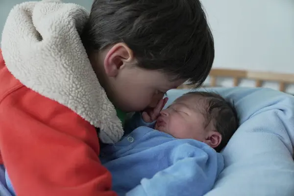 stock image A touching moment as an older child in a red jacket gently leans in to nuzzle their newborn sibling, who is wrapped in a blue onesie. The intimate moment captures the bond between siblings.