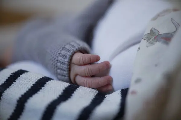 A close-up of a newborn baby\'s hand resting gently on a striped blanket. The baby is wearing a cozy gray sweater, highlighting the tenderness and delicate nature of newborn life, encapsulating warmth and innocence.