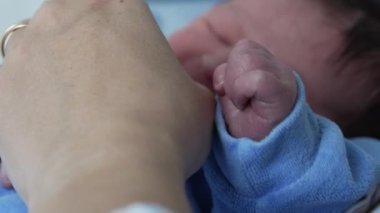 Yeni doğmuş bir bebeğin yakın plan resmi. Yumuşak mavi bir tulumun içinde uyuyor. Yüzüne yakın küçük bir eli var. Yeni doğan bebeklerin huzur ve masumiyeti bu hassas anda vurgulanıyor.