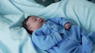 Yumuşak, koyu renk saçlı yeni doğmuş bebek, huzur içinde mavi bir tulumla uyuyor, bir hastane yatağında, yumuşak ışık ve mavi tonlarda, çocukluk döneminin masumiyetini ve sükunetini vurguluyor.