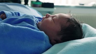 Mavi tulumlu sevimli bir bebek, bir hastane yatağında huzur içinde uyuyor, yumuşak mavi battaniyelerle çevrili, yeni hayatın özünü yakalıyor ve bir bebeğin ilk günlerinin şefkatini yakalıyor.