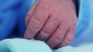 Yeni doğmuş bir bebeğin küçük elinin, yumuşak bir yüzeyde dinlenen mavi tulumundan çıkıp yeni hayatın narin ve hassas özünü yakaladığı yakın plan fotoğrafı.