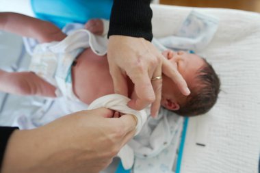 Yetişkin eller, yeni doğmuş bir bebeğe nazikçe beyaz kıyafetler giydirir, hassas bir ilgi ve ilgi anı yakalar, yeni doğmuş bir bebeği sevgi ve hassasiyetle giydirme sürecine odaklanır.