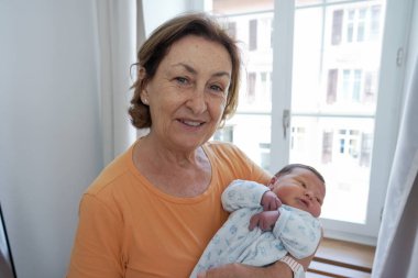 Büyükannesi yeni doğan torununu sıcak bir battaniyeye sarmış, parlak bir pencerenin önünde durmuş, aralarındaki sevgi ve bağı yakalıyor, sıcaklıkla ve ailevi gururla.