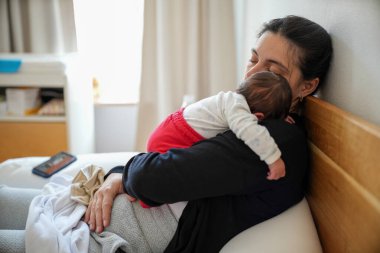 Anne, yeni doğmuş bebeği göğsünde uyurken yatakta dinleniyor. Beyaz bluz ve kırmızı pantolon giyen bebek, aralarındaki barışçıl ve hassas bağı vurgular..