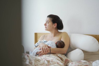 Anne, yeni doğmuş bebeğini yatakta otururken emzirirken, anne bakımı, anne bakımı ve emzirme sürecinde oluşturulan yakın bağları gösterir.