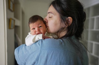 Anne nazikçe yeni doğan bebeği kucaklıyor, gözleri kapalı, bebek kameraya merakla bakarken, sevimli bir yakınlaşma anı yaratıyor kapalı bir ortamda, anne sevgisini gösteriyor.