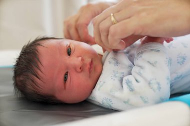 Yeni doğmuş bebek, beyaz tulum giymiş, üzerinde mavi izler var. El uzanıyor, narin parmakları ve ince deri dokusunu sergiliyor. Kapalı alanda hafif aydınlatılmış bir ortamda yakalanıyor.