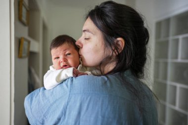 Anne yeni doğmuş bebeği sıkıca tutuyor, bebeğin başını öpüyor, bebek hafifçe açık bir ağızla ileri bakıyor, güvenli ve koşulsuz bir sevgiyi evcil bir atmosferde taşıyor.