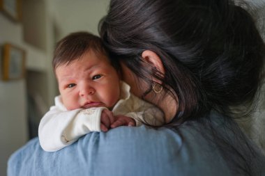 Yumuşak mavi ve beyaz giyinmiş, annesinin omzunda dinlenen yeni doğmuş bir bebek, bebek merakla etrafına bakarken, şefkat ve sevgi dolu bir anı yakalıyor.