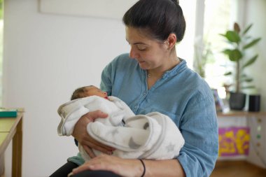 Anne, yeni doğmuş bebeğini yumuşak bir battaniyeye sarıp erken anneliğin huzurlu ve besleyici ortamını ve anne ile çocuk arasındaki derin bağı vurguluyor.