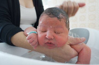 Anne yeni doğmuş bebeği beyaz bir küvette yıkıyor, bebek isim bileziği takıyor, gözleri kapalı, su yüzünden akıyor, huzurlu ve samimi bir ilgi anı ve bakım evinde erken bir bağ.