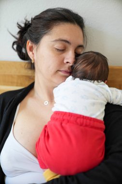 Anne ve yeni doğan bebek yatakta sakin bir anı paylaşıyorlar. Beyaz bluz ve kırmızı pantolon giyen bebek, annesinin göğsünde dinleniyor, dingin ve hassas bağı vurguluyor..